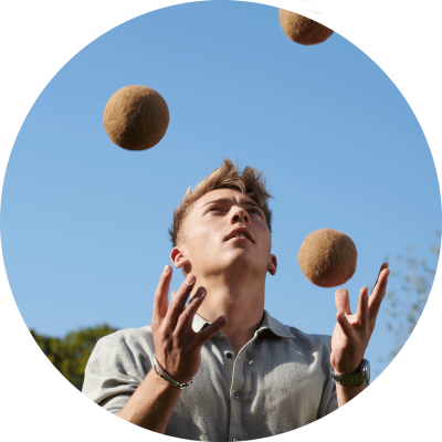 Foto rund, junger, weißer, blonder Mann jongliert mit drei braunen Bällen vor blauem Himmel